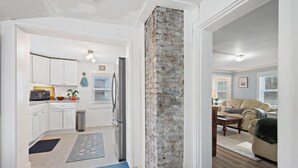 Une cuisine moderne avec des meubles blancs, un réfrigérateur et un four à micro-ondes. On y trouve un mur d’accent en briques et un espace salon avec un canapé et une table basse.