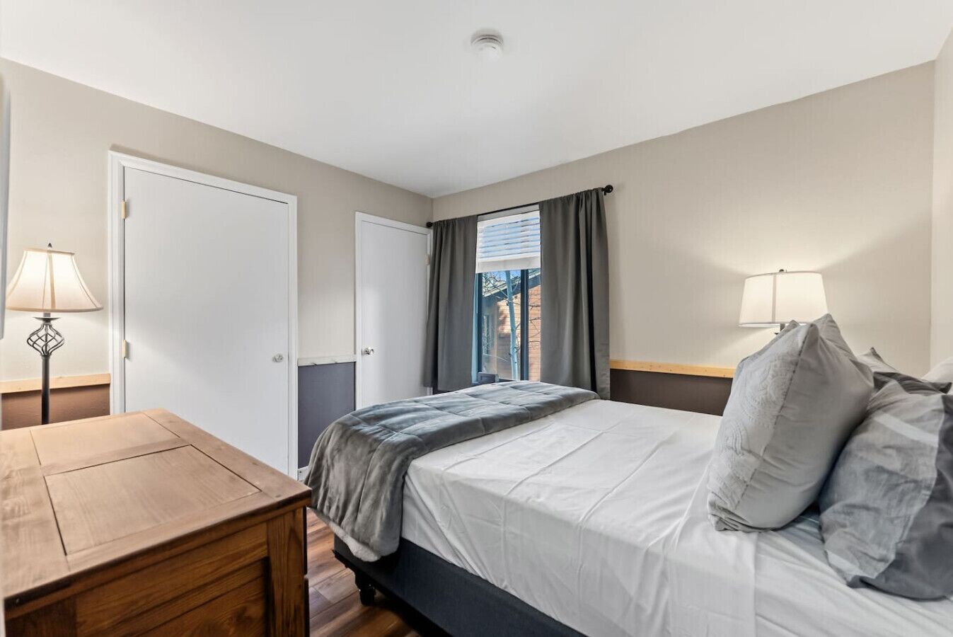 A hotel room with a bed, a wooden nightstand, a desk, and a window with curtains.