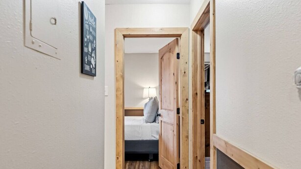 A narrow hallway with a wooden door leading to a room with a bed and a nightstand. - Mountain Charmer | Steps to Main St & Trails (Frisco)