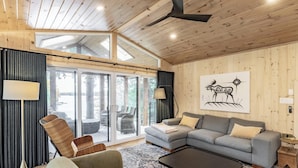 A modern living room with a sectional sofa, a floor lamp, and a large window overlooking a deck. - Delightful 4 bed 2 bath on Bass Lake (MacTier)