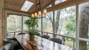 A wooden dining table with a rustic finish, surrounded by grey upholstered chairs, set in a room with large windows offering a view of trees and a forest. - Delightful 4 bed 2 bath on Bass Lake (MacTier)