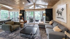 A spacious living room with a large sofa, a coffee table, and a fireplace. There are multiple windows providing natural light and a view of the outdoors. - Delightful 4 bed 2 bath on Bass Lake (MacTier)