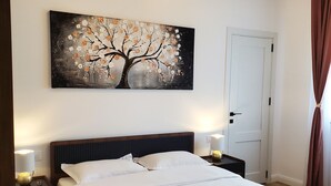 Una cama bien tendida con ropa de cama blanca y rosa, un cuadro colgado en la pared de un árbol, lámparas de noche y una puerta con cortinas.