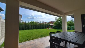 Un patio techado con mesa y sillas, un césped bien cuidado y vistas a casas al fondo.