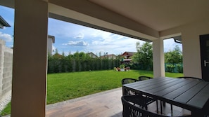 Une terrasse couverte avec une table et des chaises, une pelouse bien entretenue et une vue sur les maisons au loin.