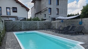 Una piscina rectangular con borde blanco, rodeada por un patio empedrado con baldosas grises. Hay sillones de descanso alineados a lo largo del borde de la piscina, y una pared de concreto con una ventana y un equipo de aire acondicionado.