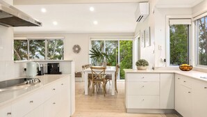 A modern kitchen with white cabinetry, a dining area with wooden chairs, and large windows offering a view of greenery. - Strandhus in Mollymoook (Mollymook Beach)