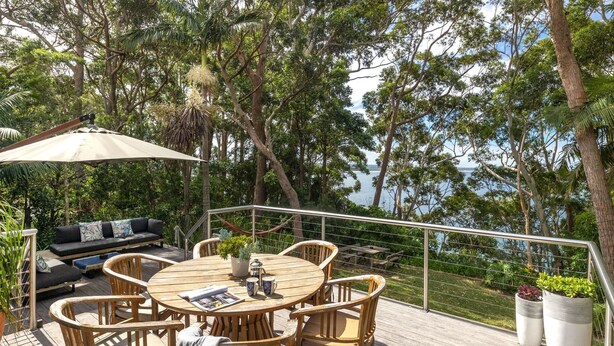 An outdoor deck with a round wooden table, wicker chairs, a sofa with cushions, and a large umbrella. - Strandhus in Mollymoook (Mollymook Beach)