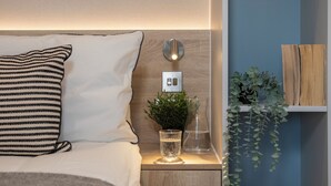 A neatly made bed with striped pillows, a bedside table with a glass of water, a small plant, and a wall-mounted light fixture. - Enclave Croydon - Campus Accommodation (Croydon)