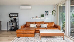 Sala de estar con sofá de cuero marrón, una mesa de centro blanca, área de cocina con electrodomésticos y vistas a través de una puerta de cristal.