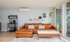 Ein Wohnzimmer mit braunem Ledersofa, einem weißen Couchtisch, einer Küchenzeile mit Geräten und Blick durch eine Glastür.