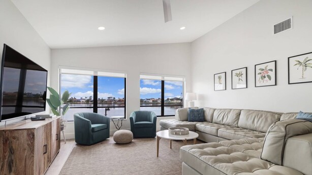 A modern living room with a large flat-screen TV, two teal armchairs, a beige sectional sofa, and a view of a lake through large windows. - 419 El Dorado Pkwy W (Cape Coral)
