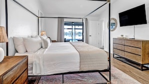 A modern bedroom with a four-poster bed, a wooden dresser, a flat-screen TV, and a view of the outdoors. - 419 El Dorado Pkwy W (Cape Coral)