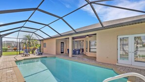 A covered swimming pool with a clear roof, a patio area with chairs, and a view of a house in the background. - 4213 SW 26th Ave (Cape Coral)