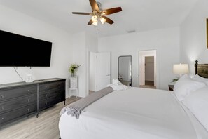 Una camera da letto con un letto, una cassettiera, una ventola a soffitto, una televisione, uno specchio e una porta che dà su un'altra stanza.