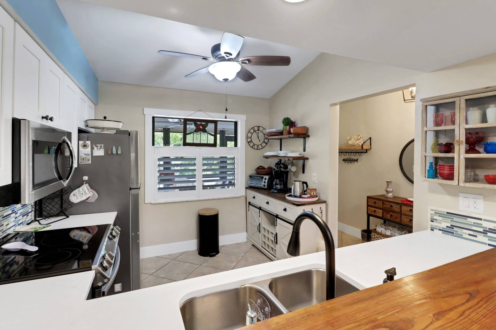 Una cocina moderna con horno empotrado, microondas y pileta de acero inoxidable. Hay un ventilador de techo, una ventana con persianas y un reloj en la pared.