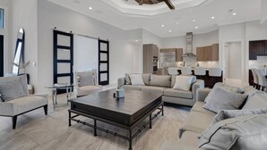 A spacious living room with a large dark wood coffee table, a sofa set, and a kitchen area with modern appliances. - 2301 SW 39th St (Cape Coral)