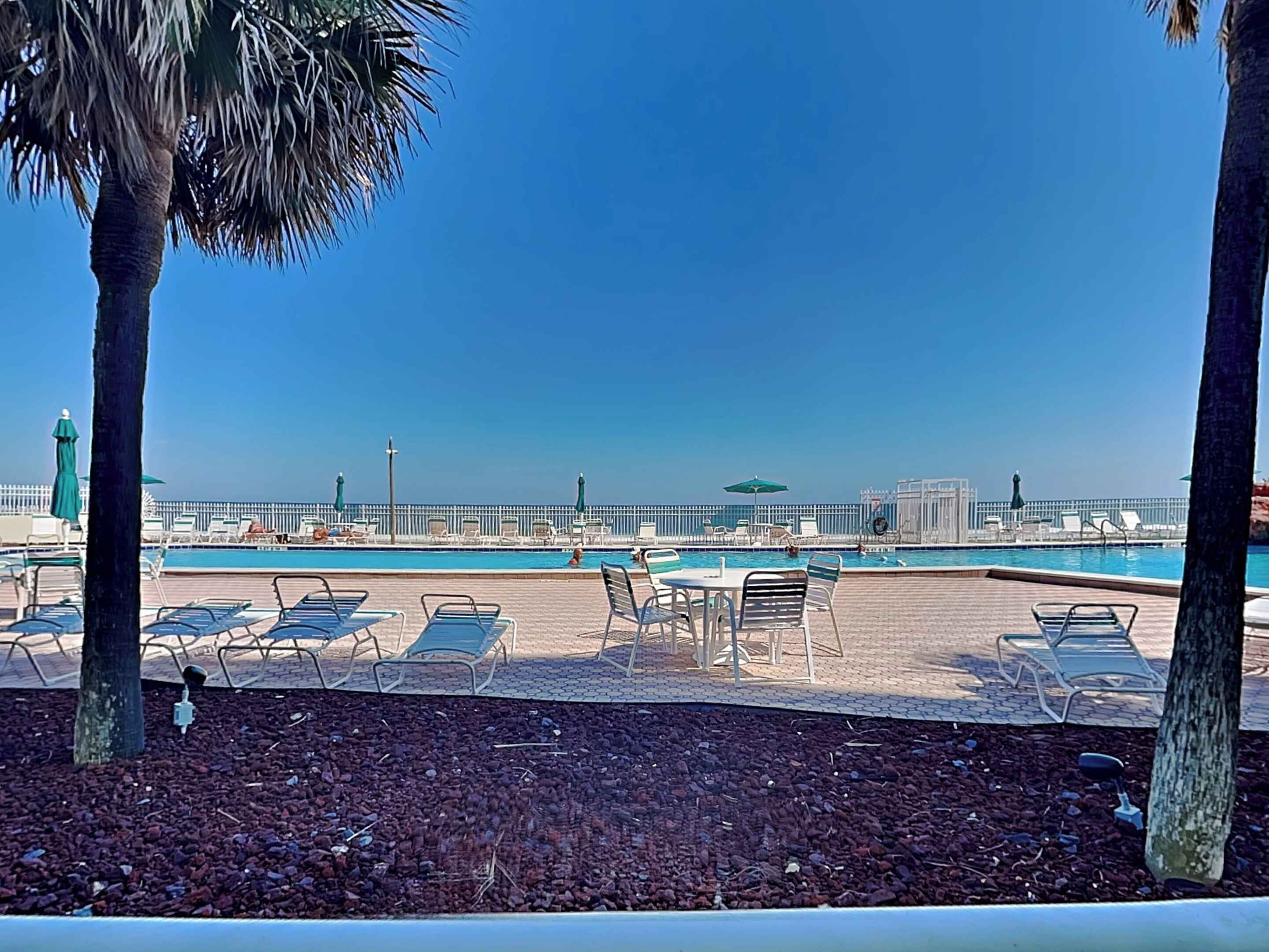 A poolside area with lounge chairs and umbrellas, a clear sky, and a distant cityscape.