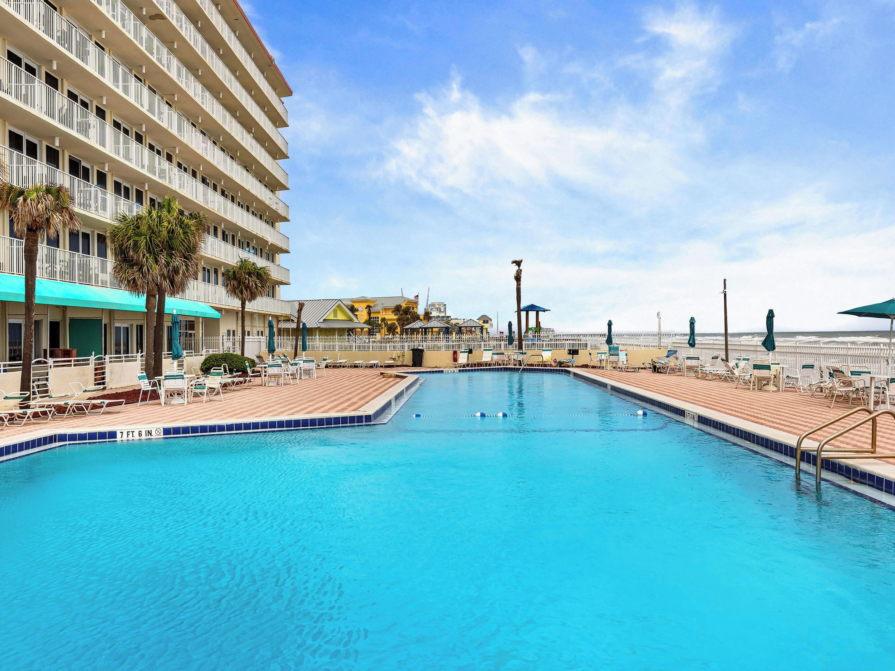 A swimming pool with lounge chairs and umbrellas, a beach in the background, and a hotel building with balconies.