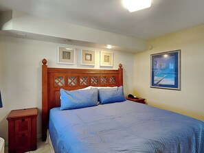 Een slaapkamer met een houten hoofdbord, een groot bed met blauw beddengoed, een nachtkastje, een ingelijst kunstwerk aan de muur en een schilderij van een zwembad.