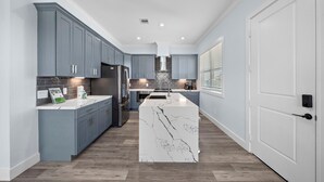A modern kitchen with grey cabinets, a central island, and a stainless steel refrigerator. - Beachside Serenity (Galveston)