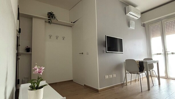 A modern living room with a flat-screen TV mounted on the wall, a dining area with a table and chairs, and a small white cabinet with a potted orchid. - Civic8-Navigli-Iulm (Milano)