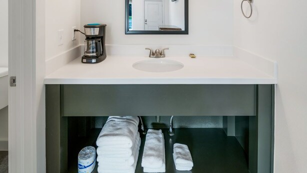 A modern bathroom with a white vanity, a large mirror, and a built-in sink. There is a towel rack above the sink and a towel dispenser below. - Affordable Lodge w/Covered, Lighted, Pull Through Parking. Grills and Outdoor TV (Grove)