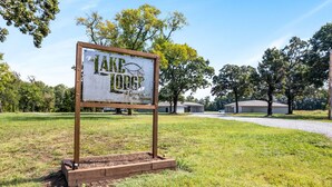 A sign for "Lake Lodge" with a wooden frame and metal letters, set in a grassy area with trees and buildings in the background. - Affordable Lodge w/Covered, Lighted, Pull Through Parking. Grills and Outdoor TV (Grove)