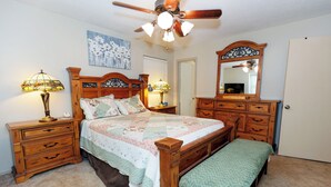 A bedroom with a wooden bed, two bedside tables with lamps, a bench, a ceiling fan, a dresser, and a mirror.