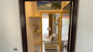 A hallway leading to a bedroom with a bed, a nightstand, and a lamp. There is a small table and a chair in the corner, and a door with a handle.