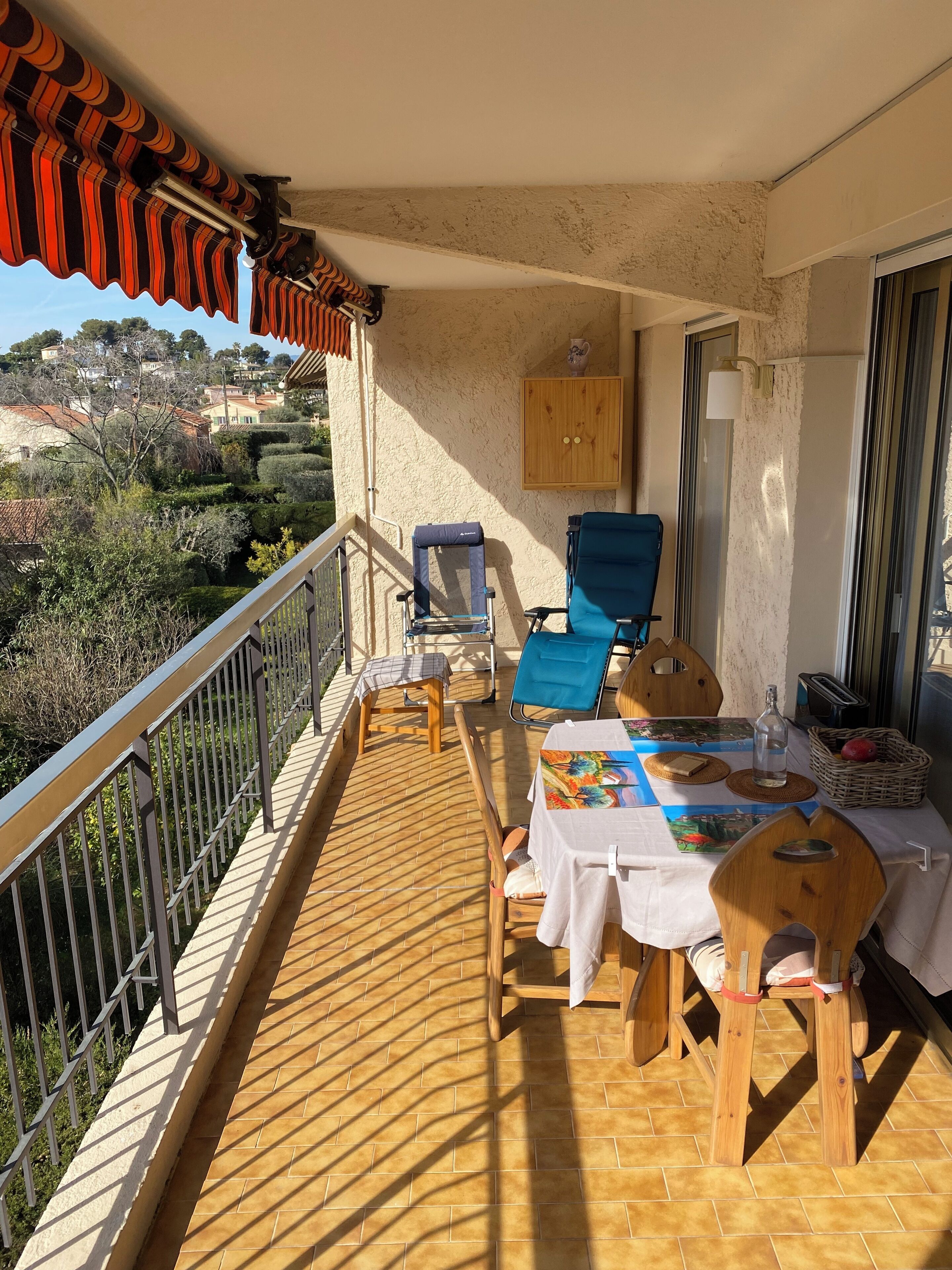 Un balcón con mesa lista para comer, una silla azul, una silla de madera y una mesita con una botella y una cesta de fruta.