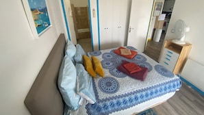 A bedroom with a bed, pillows, a bedside table with a fan, a mirror, and a door leading to a bathroom.