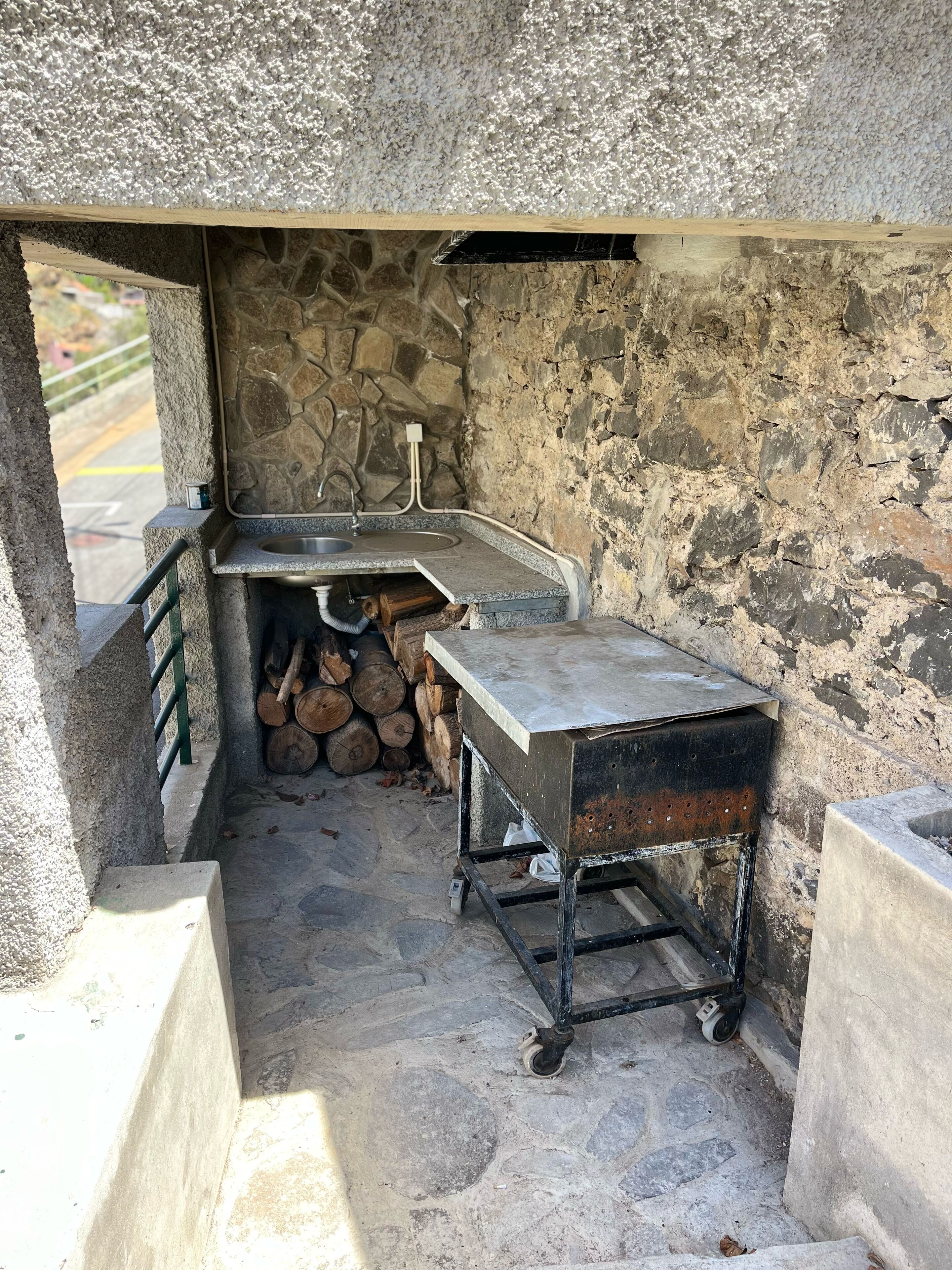 Une cuisine extérieure avec des murs en pierre, équipée d’un évier, d’une table en métal et d’une pile de bois de chauffage.