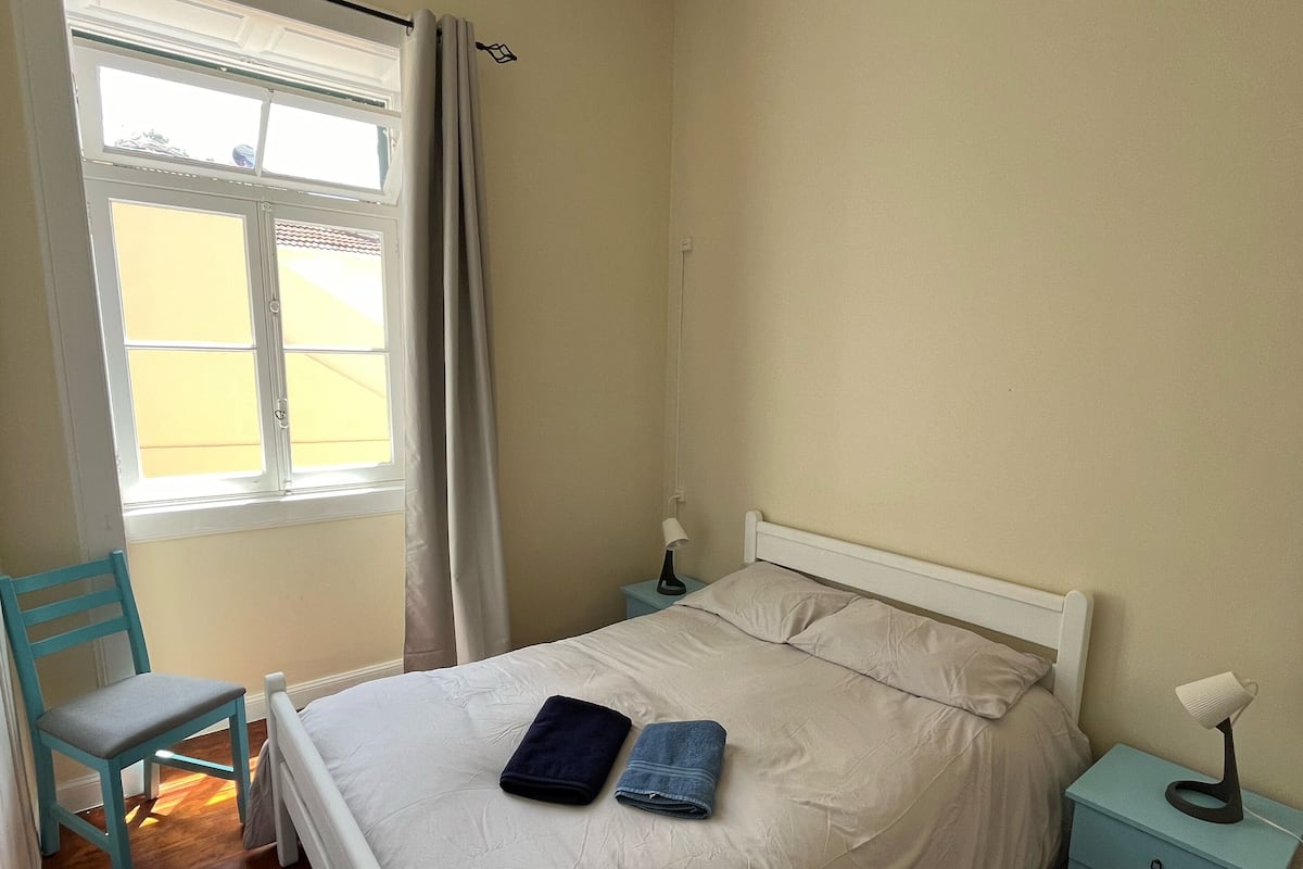 A neatly made bed with two folded towels, a nightstand with a lamp, a chair, and a window with curtains.