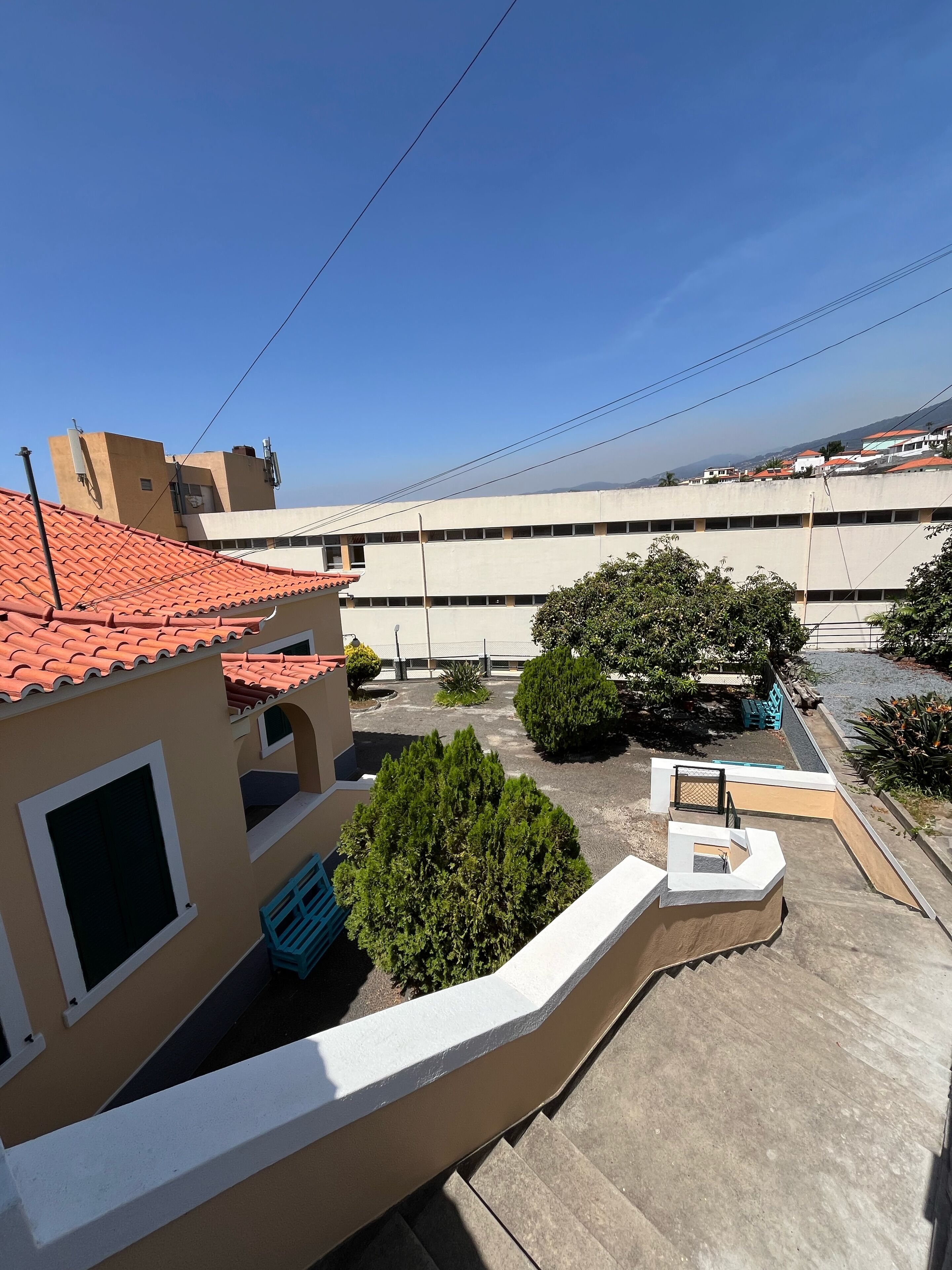 Um bairro residencial com um edifício de telhado vermelho, um pequeno jardim com uma árvore e um espaço pavimentado com bancos.