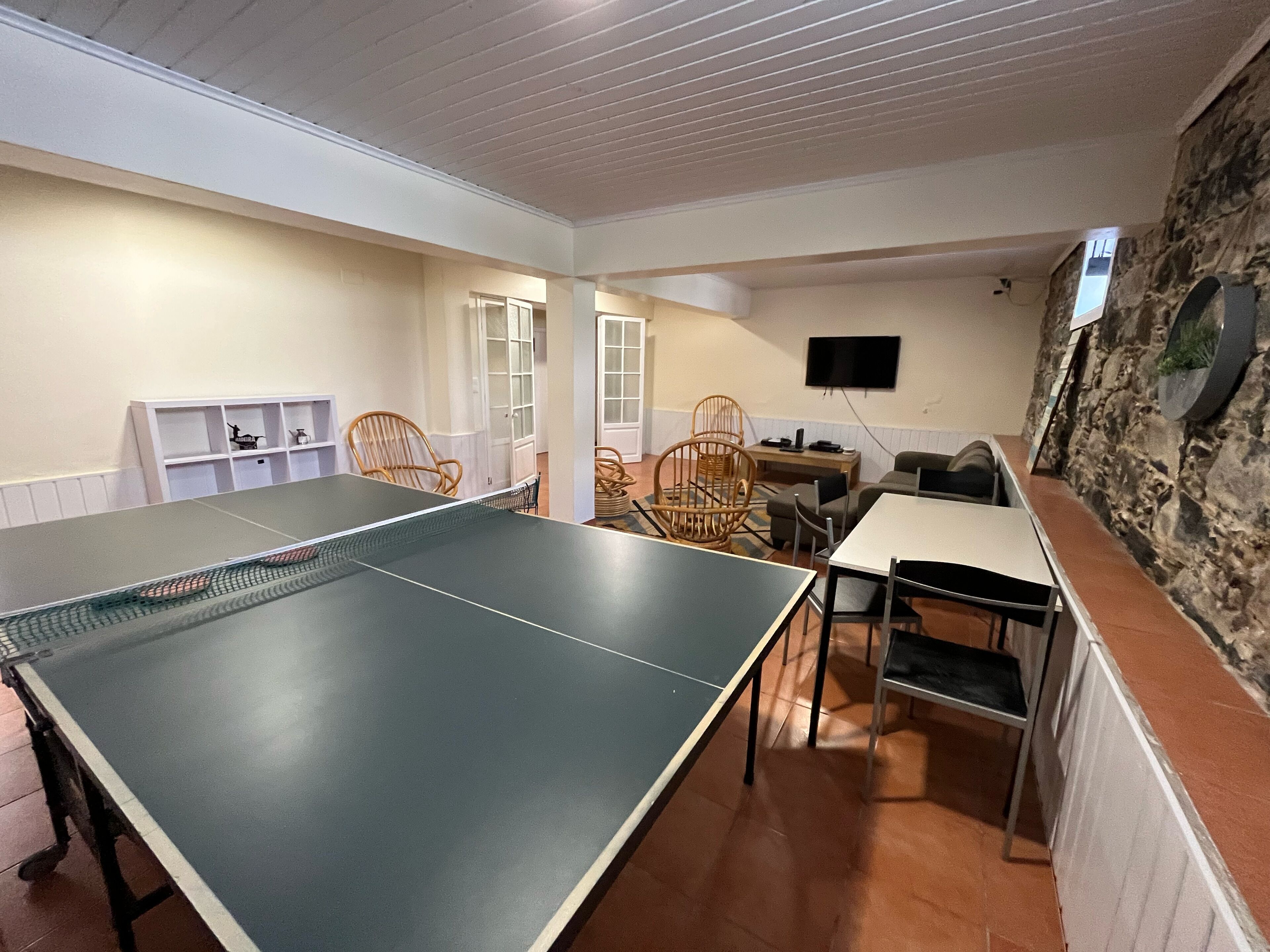 A room with a ping pong table, wicker chairs, a small table with a TV, and a stone wall.