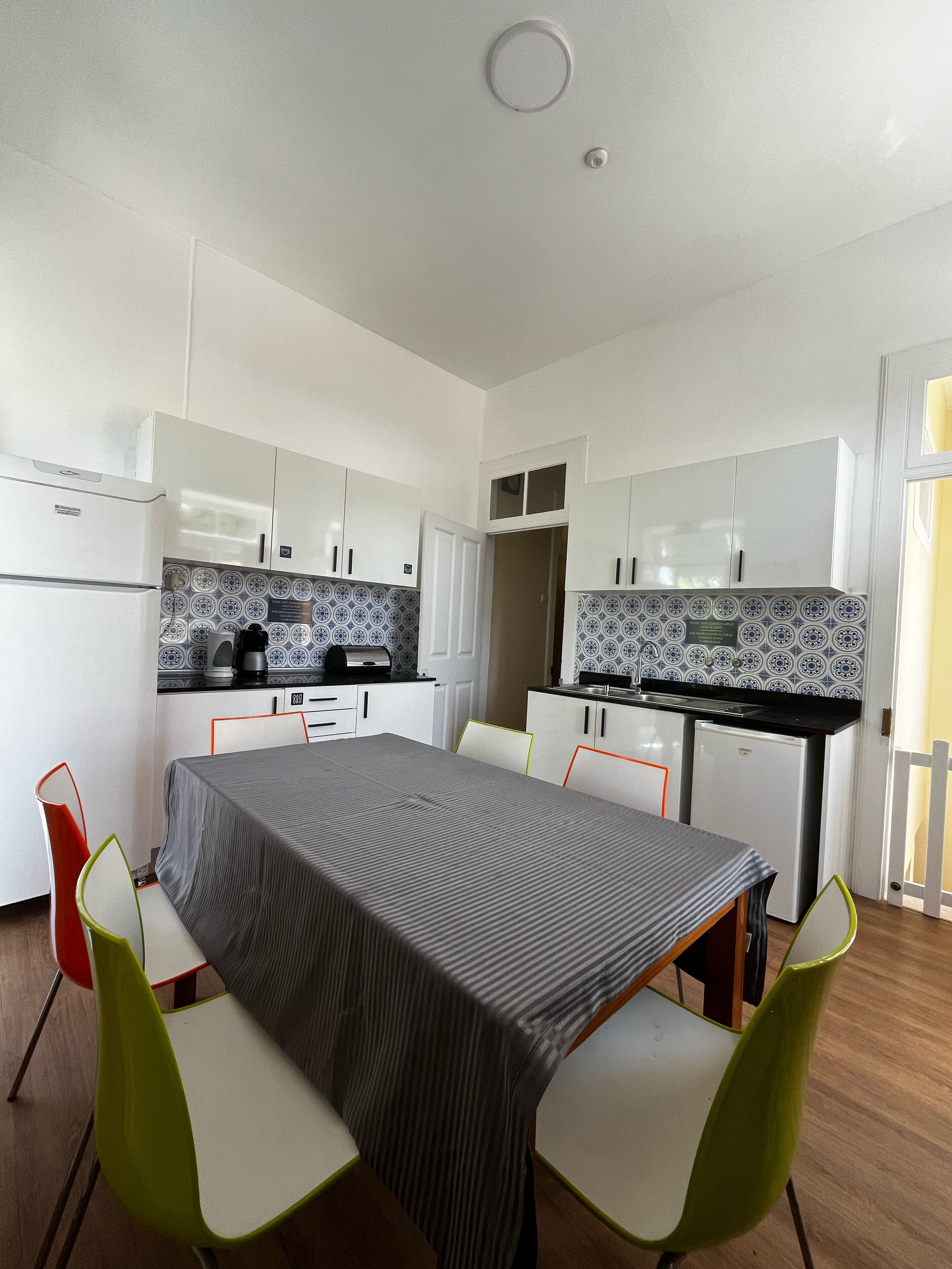 A compact kitchen with white cabinets, a small refrigerator, and a coffee maker. There is a dining table with a striped tablecloth and two chairs.