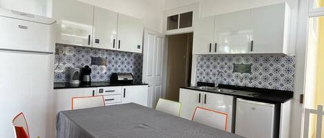 A compact kitchen with white cabinets, a small refrigerator, and a coffee maker. There is a dining table with a striped tablecloth and two chairs.