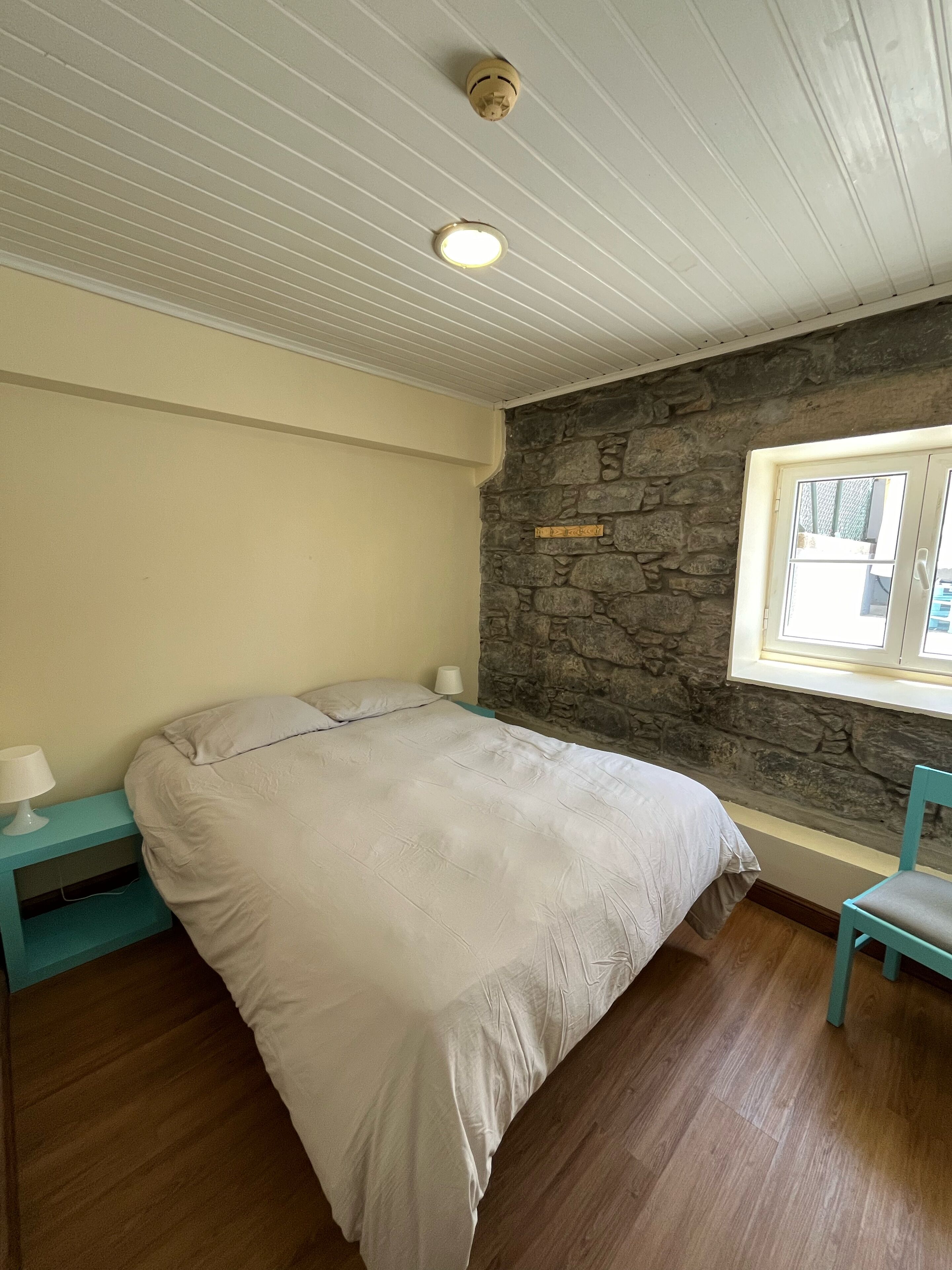 Un dormitorio con cama, mesita de noche, pared de piedra, ventana y una silla.