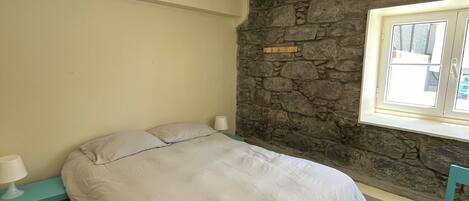 Un dormitorio con cama, mesita de noche, pared de piedra, ventana y una silla.