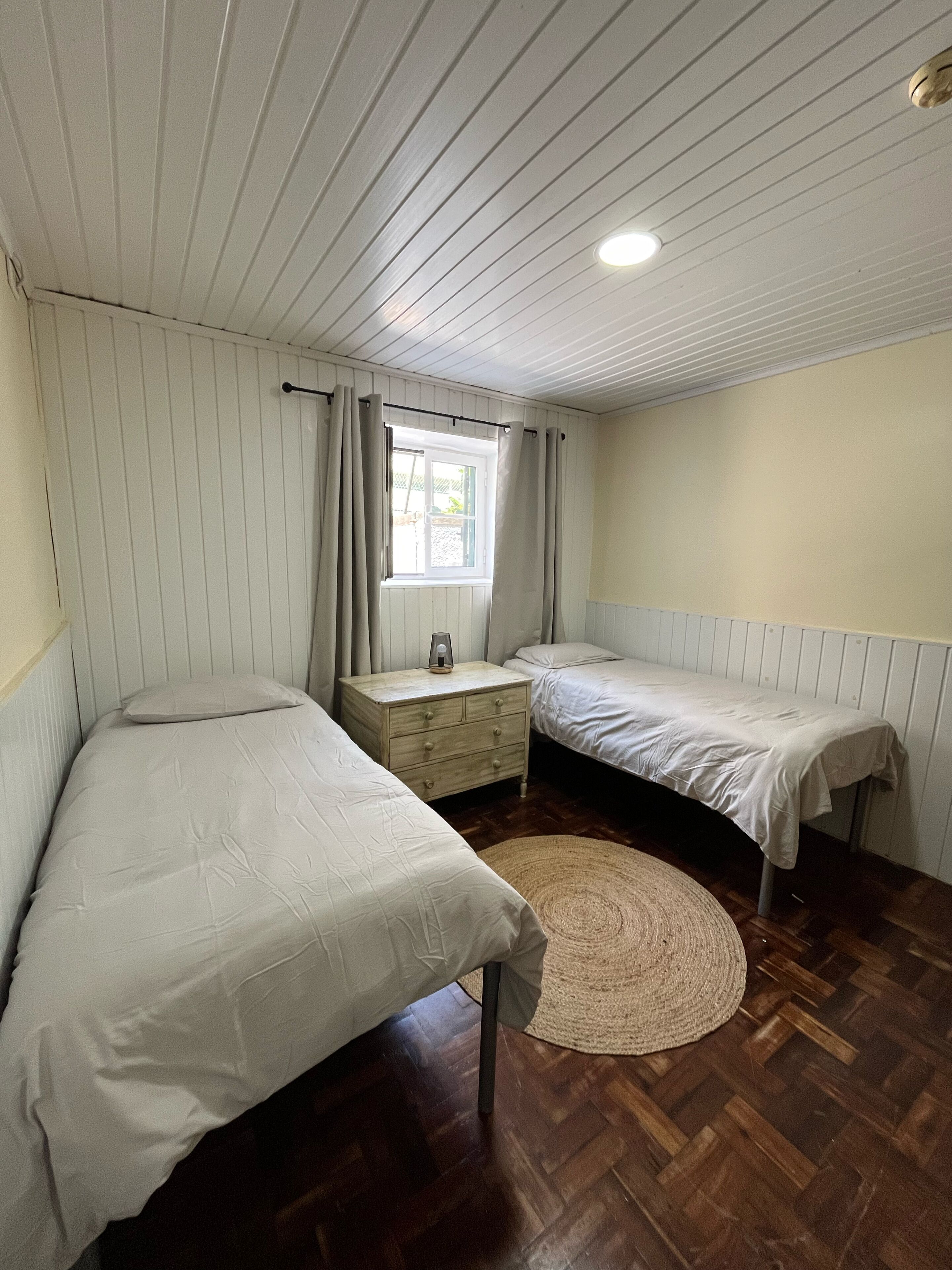 Een kleine, eenpersoonskamer met twee eenpersoonsbedden, een houten nachtkastje, een raam met gordijnen en een geweven vloerkleed op een houten vloer.