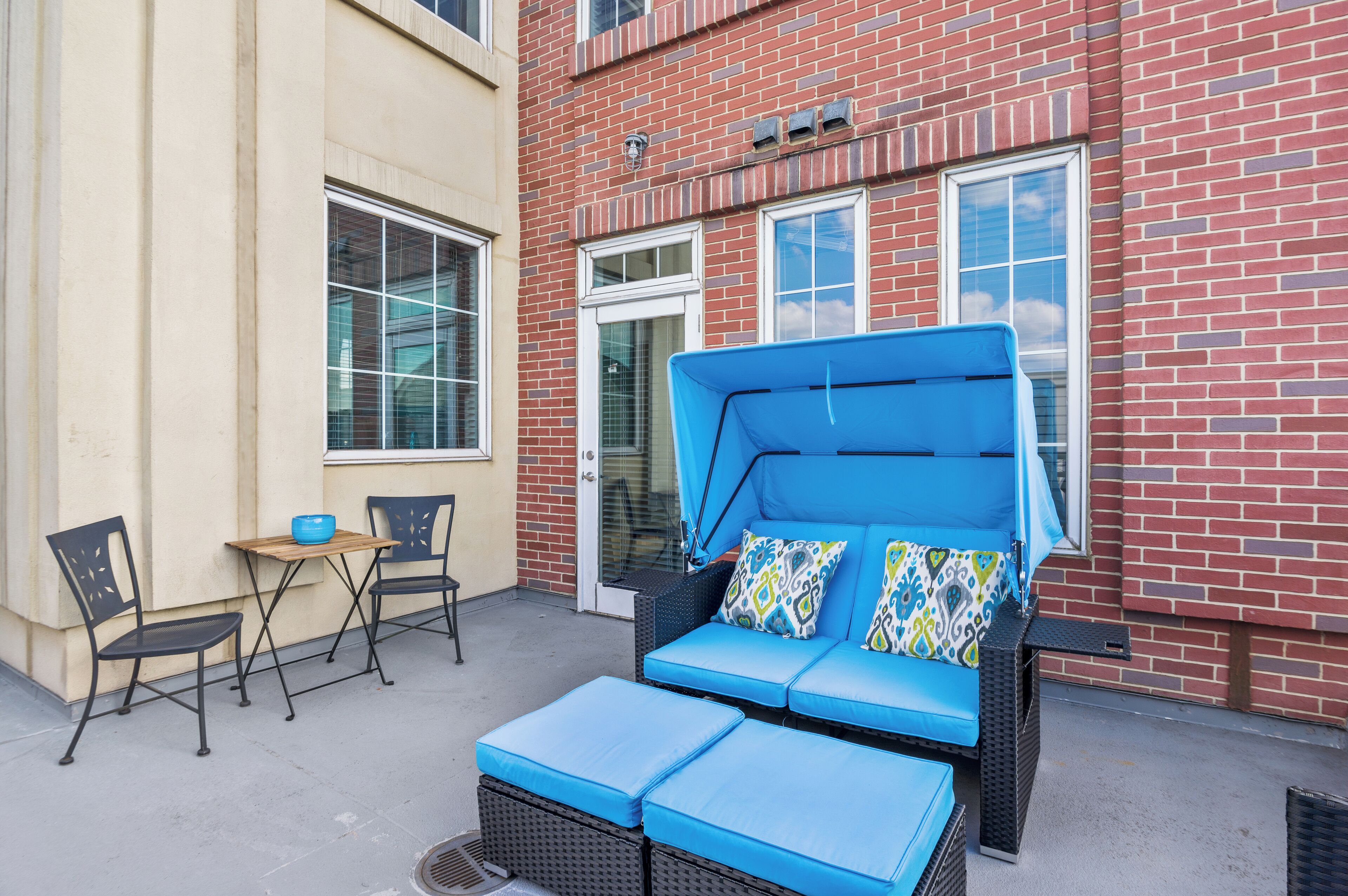 An outdoor seating area with a blue sofa and chairs, a small table, and a window with blinds.