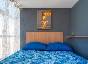 A bedroom with a bed, a headboard, a wall-mounted giraffe painting, and a window with patterned curtains.