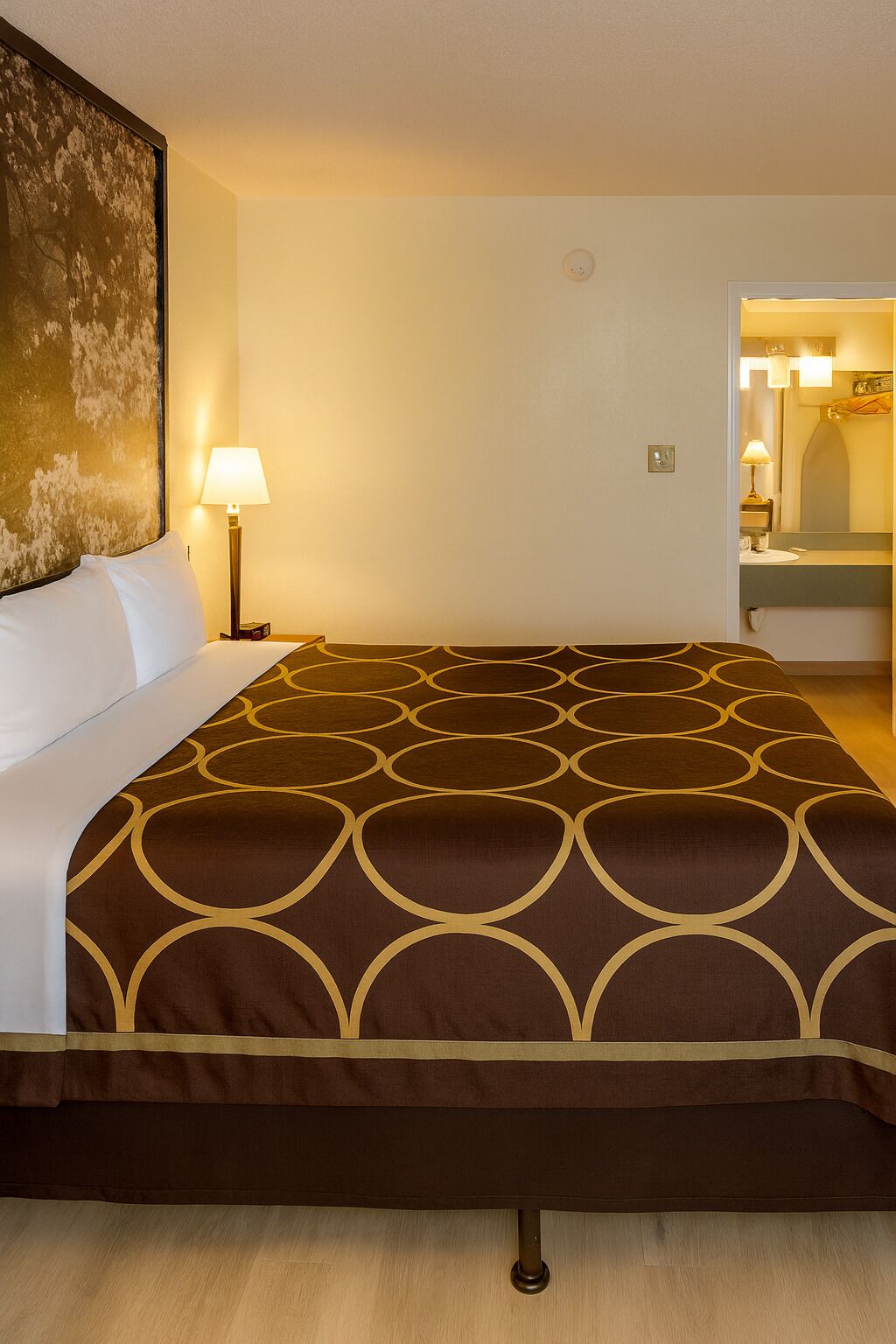 Una habitación de hotel con una cama grande, una mesita de noche con una lámpara, un cuadro en la pared y un baño visible a través de una puerta abierta.