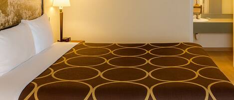 Una habitación de hotel con una cama grande, una mesita de noche con una lámpara, un cuadro en la pared y un baño visible a través de una puerta abierta.