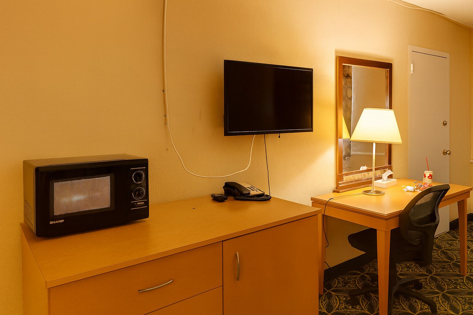 A hotel room with a desk, a television, a telephone, a lamp, and a mirror.