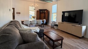 Ein modernes Wohnzimmer mit einem Ecksofa, einem Holztisch, einem Fernseher auf einer Holzkommode und einem Essbereich mit einer Pendelleuchte.