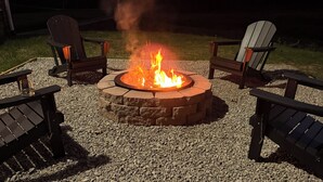 Ein Lagerfeuer mit vier Adirondack-Stühlen darum herum, auf einer Schotterfläche mit Rasen und Bäumen im Hintergrund.
