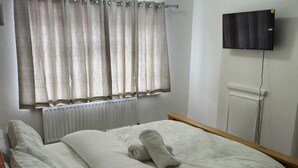 Ein Schlafzimmer mit einem Bett, einem Heizkörper, einem Fenster mit Vorhängen und einem an der Wand befestigten Fernseher.