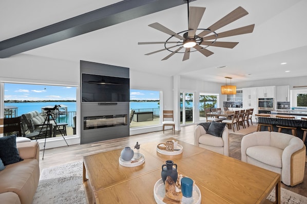 A spacious living area with a large window overlooking a body of water, a ceiling fan, and a modern kitchen with white cabinets.
